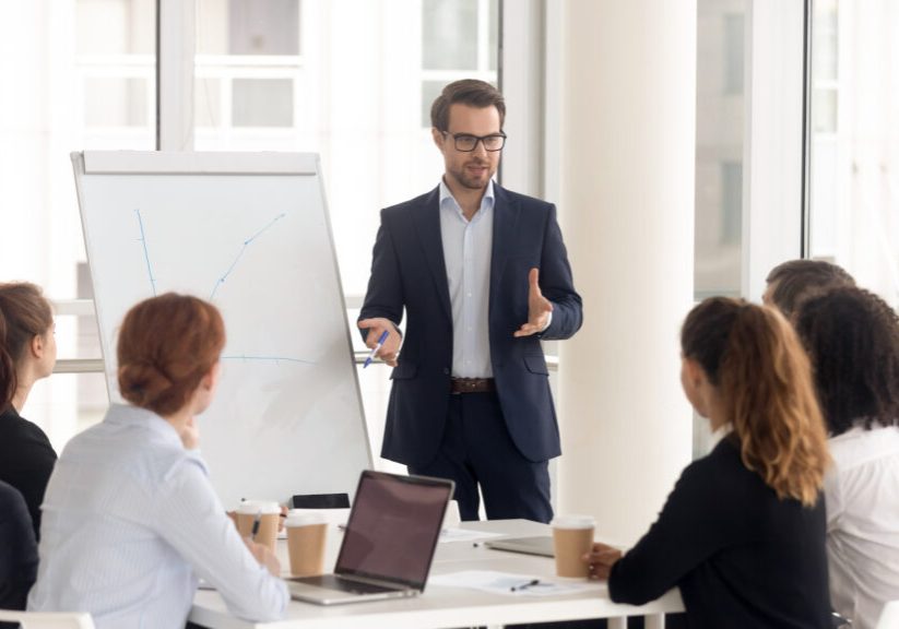 Male business coach speaker in suit give flipchart presentation, speaker presenter consulting training persuading employees client group, mentor leader explain graph strategy at team meeting workshop